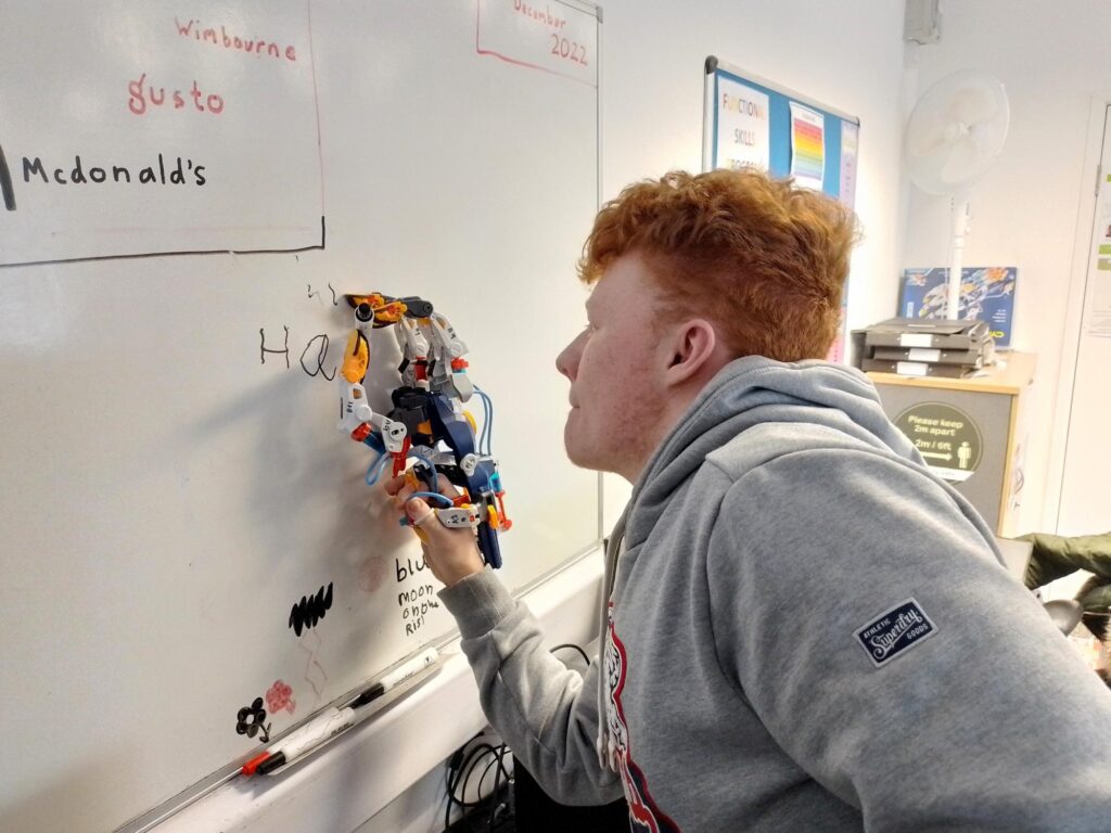 One student playing with a robot toy holding a pen using a pen on a whiteboard in a classroom at the EMA Moreton Campus.